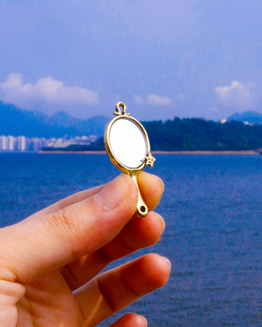 hand holding a brass miniature tiny hand mirror, featuring engraved star motifs on both sides and a functional reflective mirror surface, shown as wearable charm