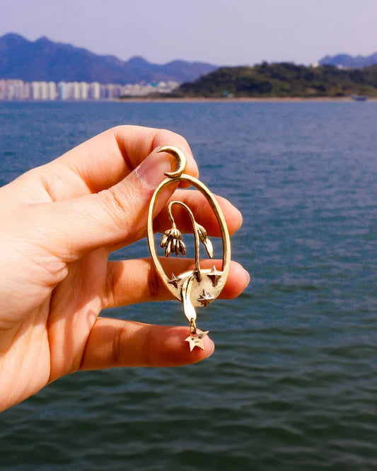 Bloom of a Thousand Stars’ handmade oval earring featuring a dying flower, crescent moon, and falling leaf with dangling star. Lost-wax cast in brass—a wearable poem about grief and cosmic rebirth