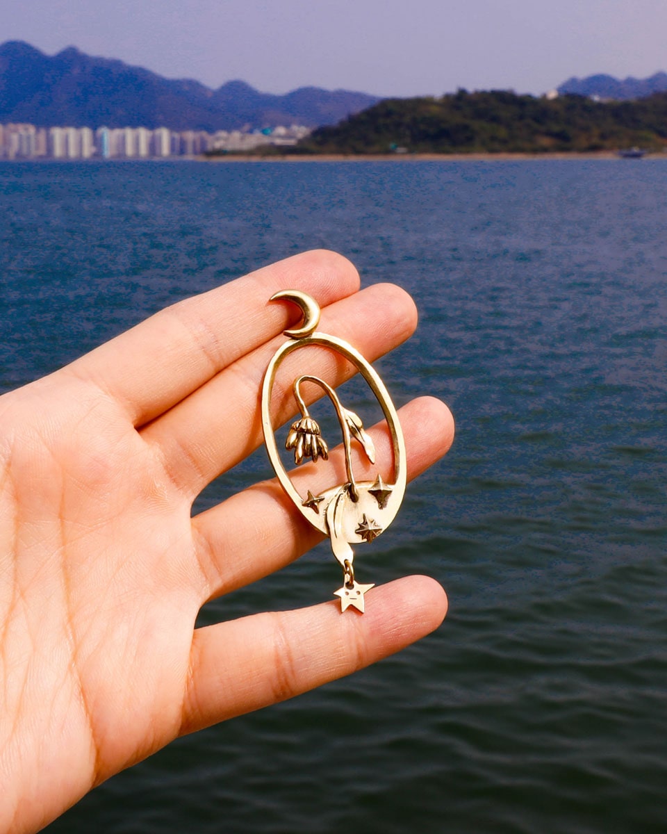 Bloom of a Thousand Stars’ handmade oval earring featuring a dying flower, crescent moon, and falling leaf with dangling star. Lost-wax cast in brass—a wearable poem about grief and cosmic rebirth