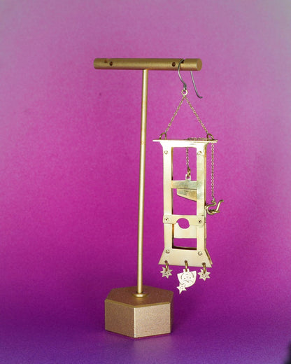 Handmade brass guillotine earring with a movable blade, chain mechanism, dangling stars, and a suspended face inspired by the Person Controlled by the Stars series.