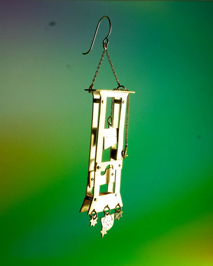 Handmade brass guillotine earring with a movable blade, chain mechanism, dangling stars, and a suspended face inspired by the Person Controlled by the Stars series.
