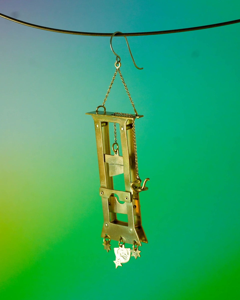 Handmade brass guillotine earring with a movable blade, chain mechanism, dangling stars, and a suspended face inspired by the Person Controlled by the Stars series.