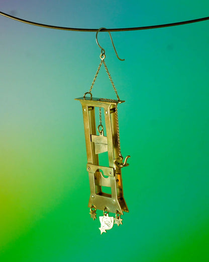 Handmade brass guillotine earring with a movable blade, chain mechanism, dangling stars, and a suspended face inspired by the Person Controlled by the Stars series.
