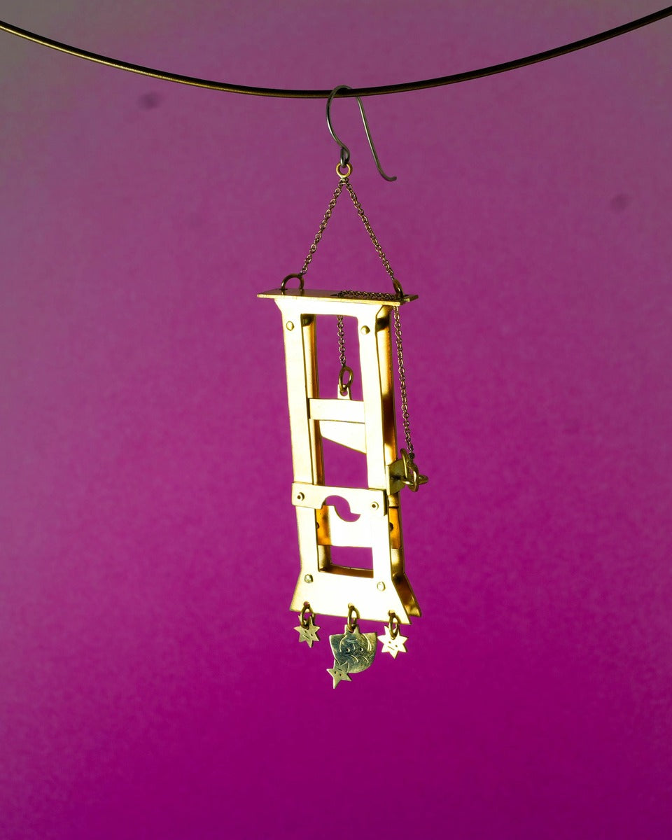 Handmade brass guillotine earring with a movable blade, chain mechanism, dangling stars, and a suspended face inspired by the Person Controlled by the Stars series.