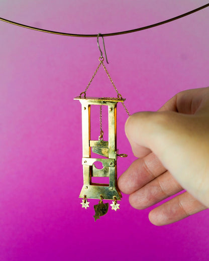 Small guillotine model held by a hand, Handmade brass guillotine earring with a movable blade, chain mechanism, dangling stars, and a suspended face inspired by the Person Controlled by the Stars series.