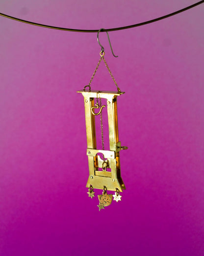 Handmade brass guillotine earring with a movable blade, chain mechanism, dangling stars, and a suspended face inspired by the Person Controlled by the Stars series.