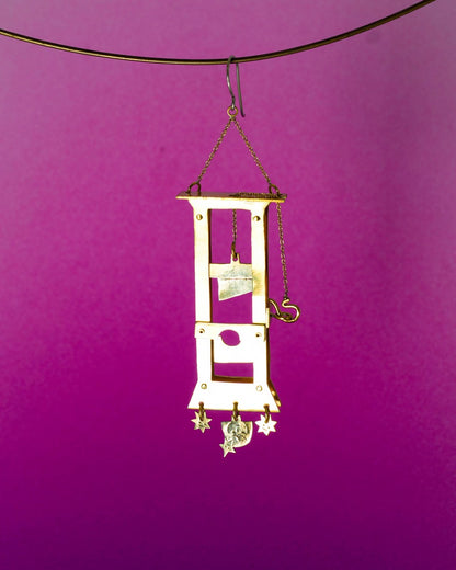 Handmade brass guillotine earring with a movable blade, chain mechanism, dangling stars, and a suspended face inspired by the Person Controlled by the Stars series.