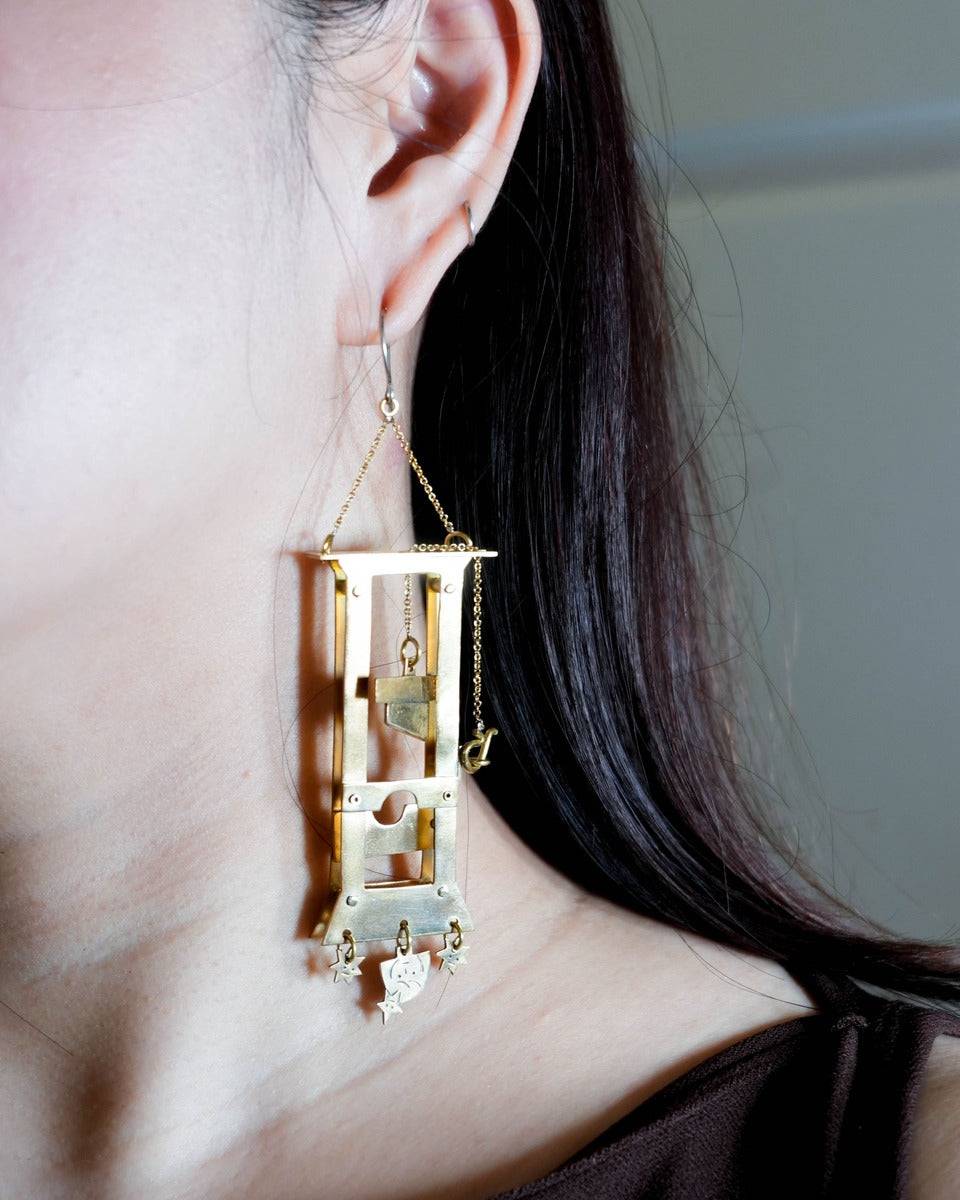 person wearing a handmade brass guillotine earring with a movable blade, chain mechanism, dangling stars, and a suspended face inspired by the Person Controlled by the Stars series.