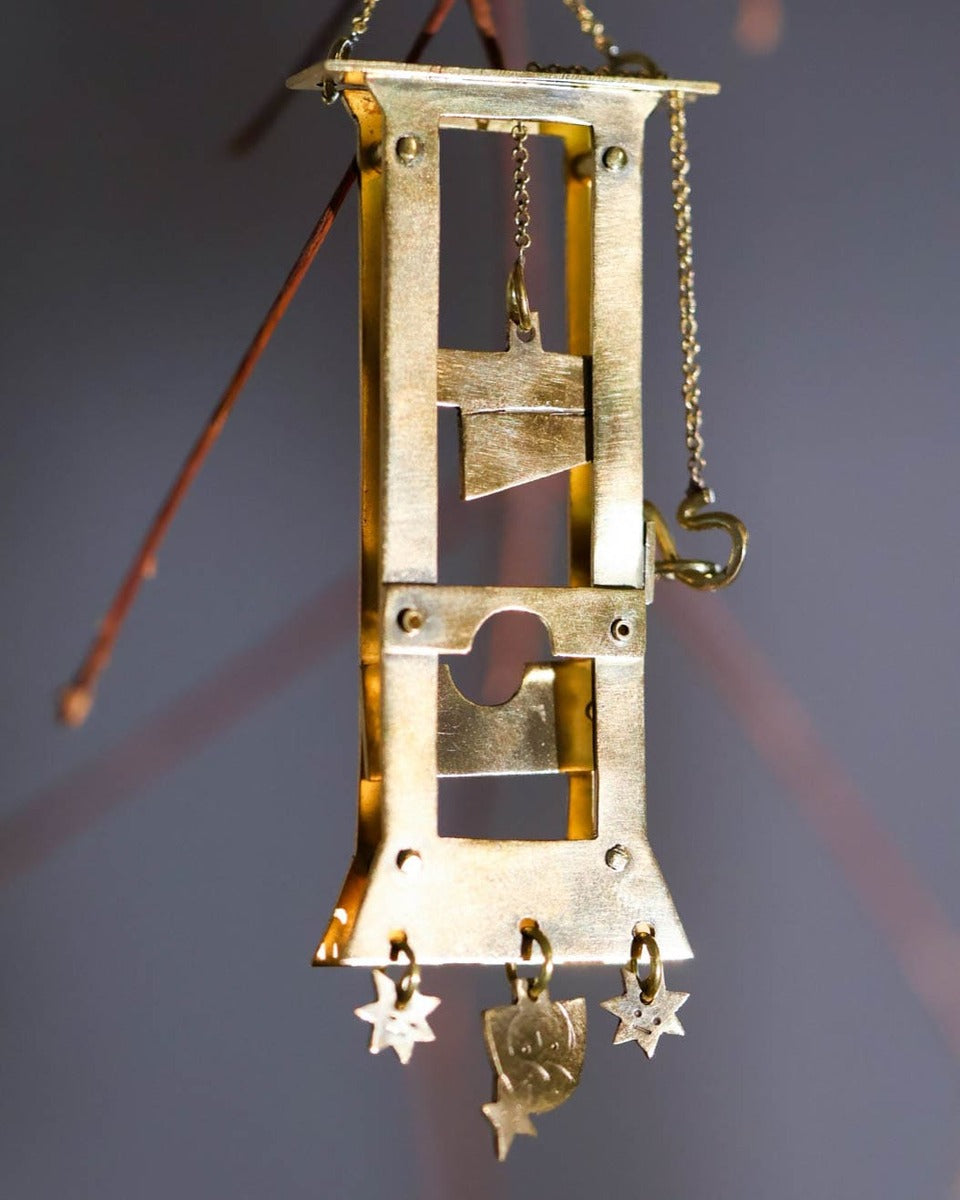 Handmade brass guillotine earring with a movable blade, chain mechanism, dangling stars, and a suspended face inspired by the Person Controlled by the Stars series.
