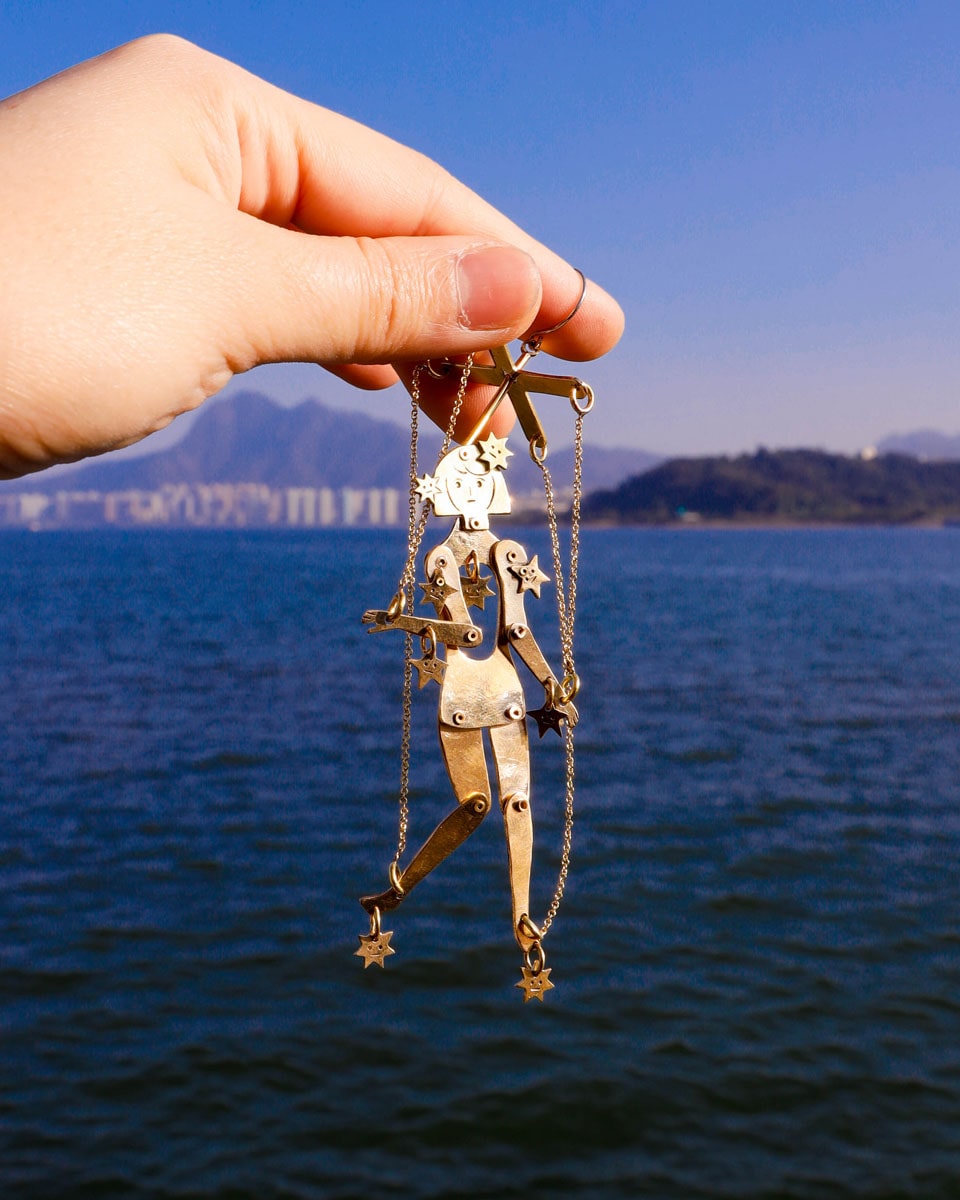 hand holding an articulated brass puppet earring "The Person Controlled by the Stars" with moving handsaw joints