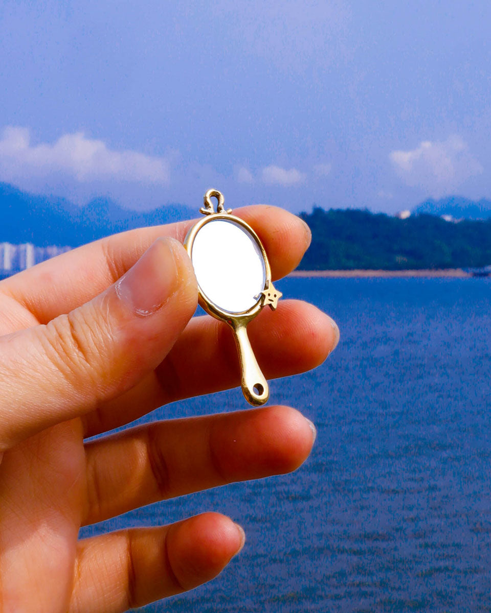 hand holding a brass miniature tiny hand mirror, featuring engraved star motifs on both sides and a functional reflective mirror surface, shown as wearable charm 