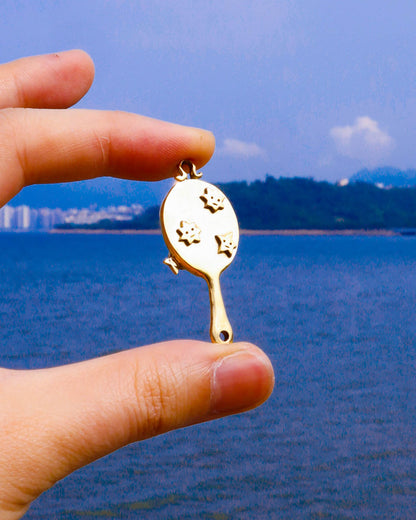 hand holding a brass miniature tiny hand mirror, featuring engraved star motifs on both sides and a functional reflective mirror surface, shown as wearable charm 