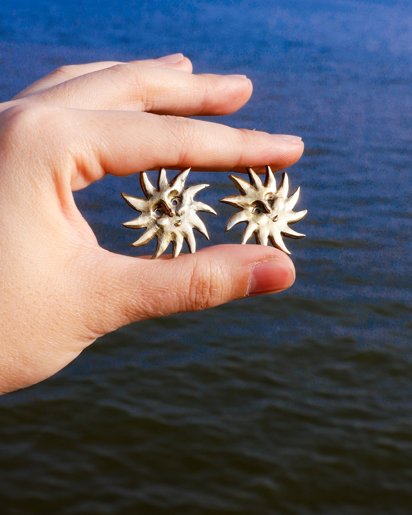 Hand holding two sun-shaped earrings with a blurred water background