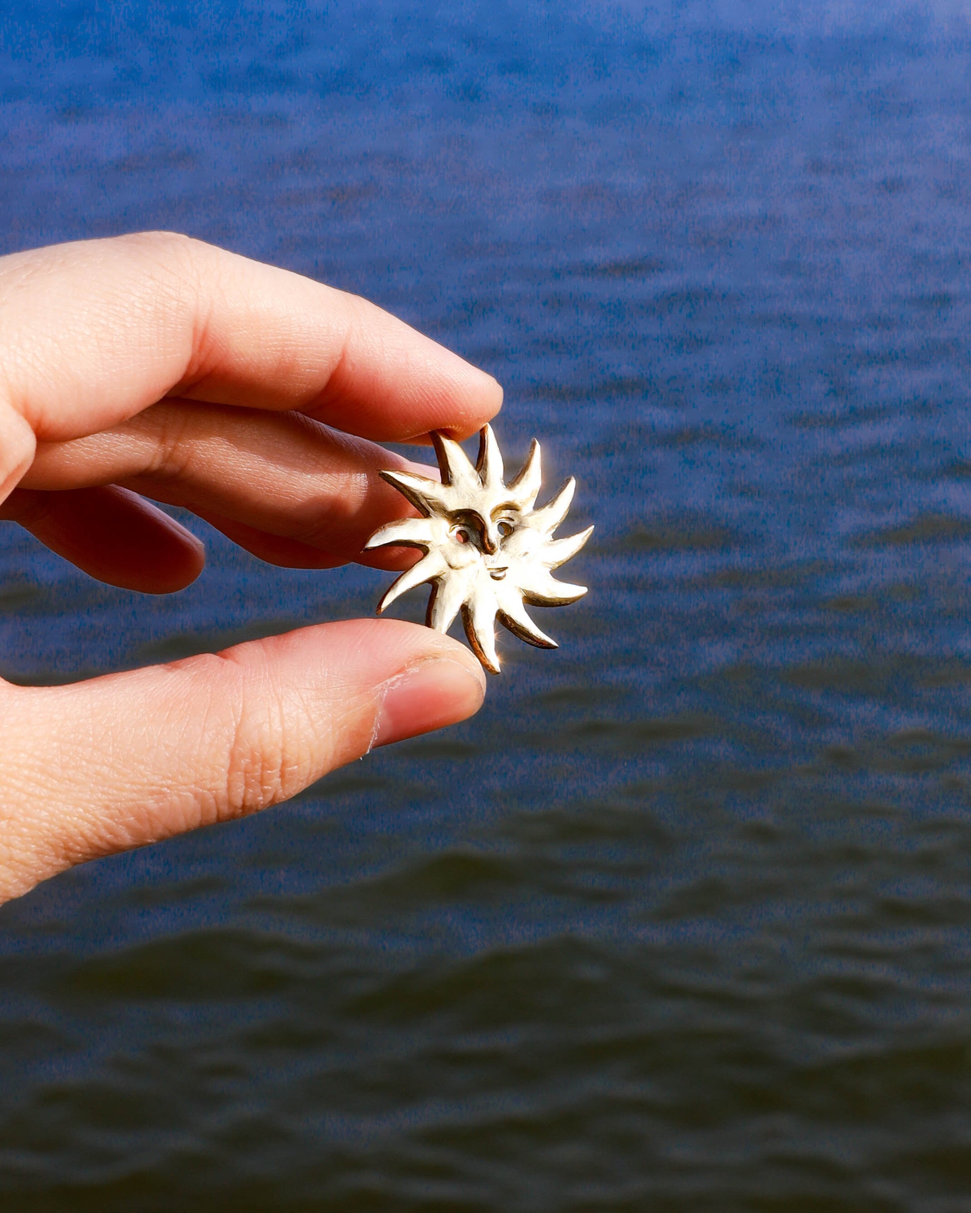 Hand holding a gold sun-shaped earring against a blue water background